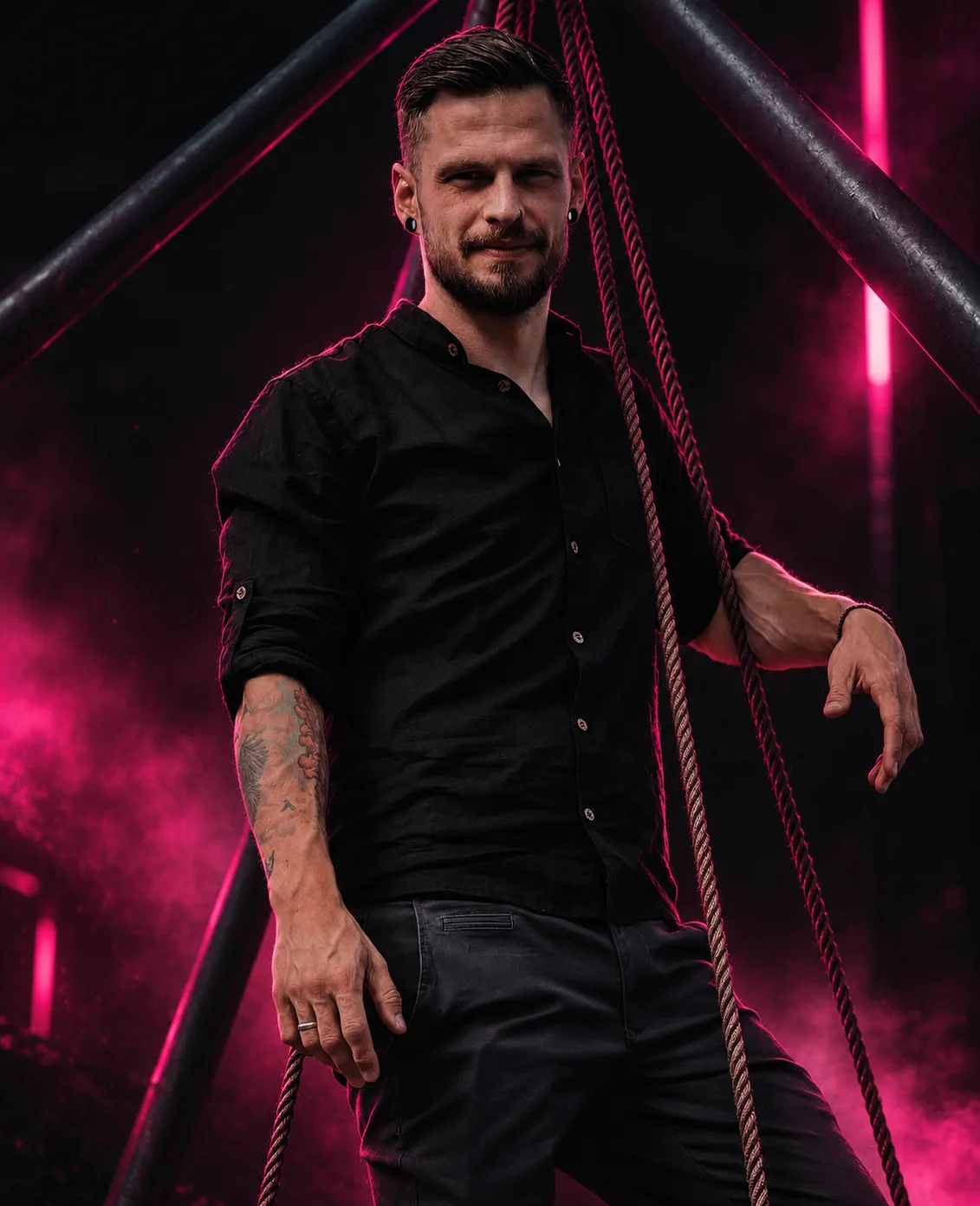 Alex in a black shirt, leaning against a rope between steel beams in pink light